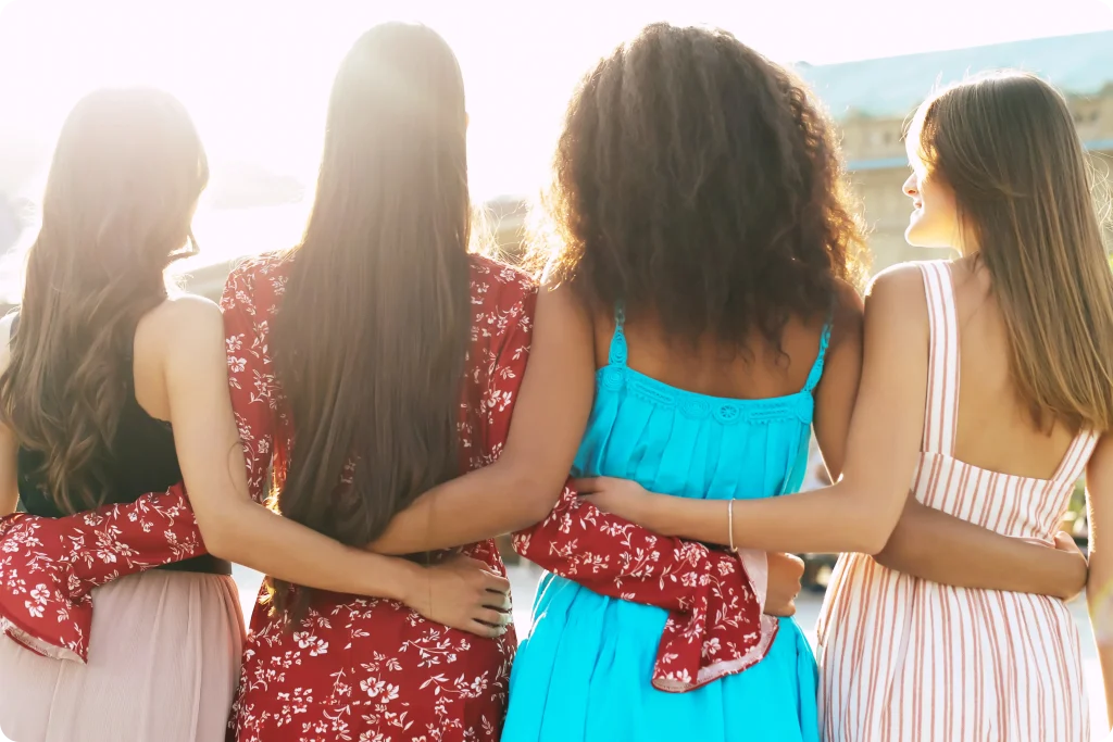 Four women stand arm in arm, seen from behind, enjoying warm sunlight outdoors, showing friendship, diversity, and support together unity.